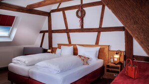 A room with a wooden ceiling, a bed with white bedding, a red upholstered bench, a wooden nightstand with a lamp, and a window with blinds.