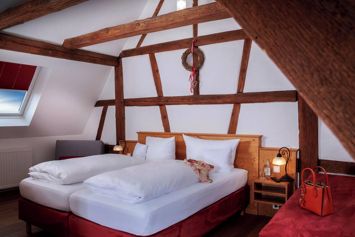 A room with a wooden ceiling, a bed with white bedding, a red upholstered bench, a wooden nightstand with a lamp, and a window with blinds.