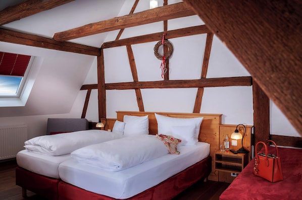Una habitación con techo de madera, una cama con ropa blanca, un banco tapizado en rojo, una mesita de noche de madera con una lámpara y una ventana con persianas.