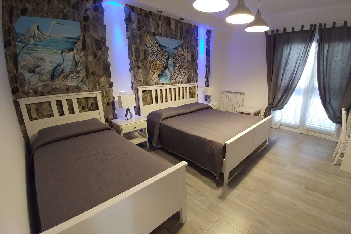 Hotellihuone, jossa on kaksi sänkyä, kiviseinästä tehty seinämaalaus, ruokailutila pöytineen ja tuoleineen sekä ikkuna, jossa on verhot.