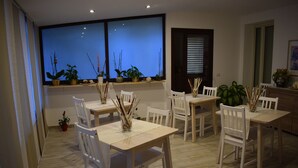 Un área de comedor con sillas y mesas blancas, un ventanal con plantas y una puerta con persianas.
