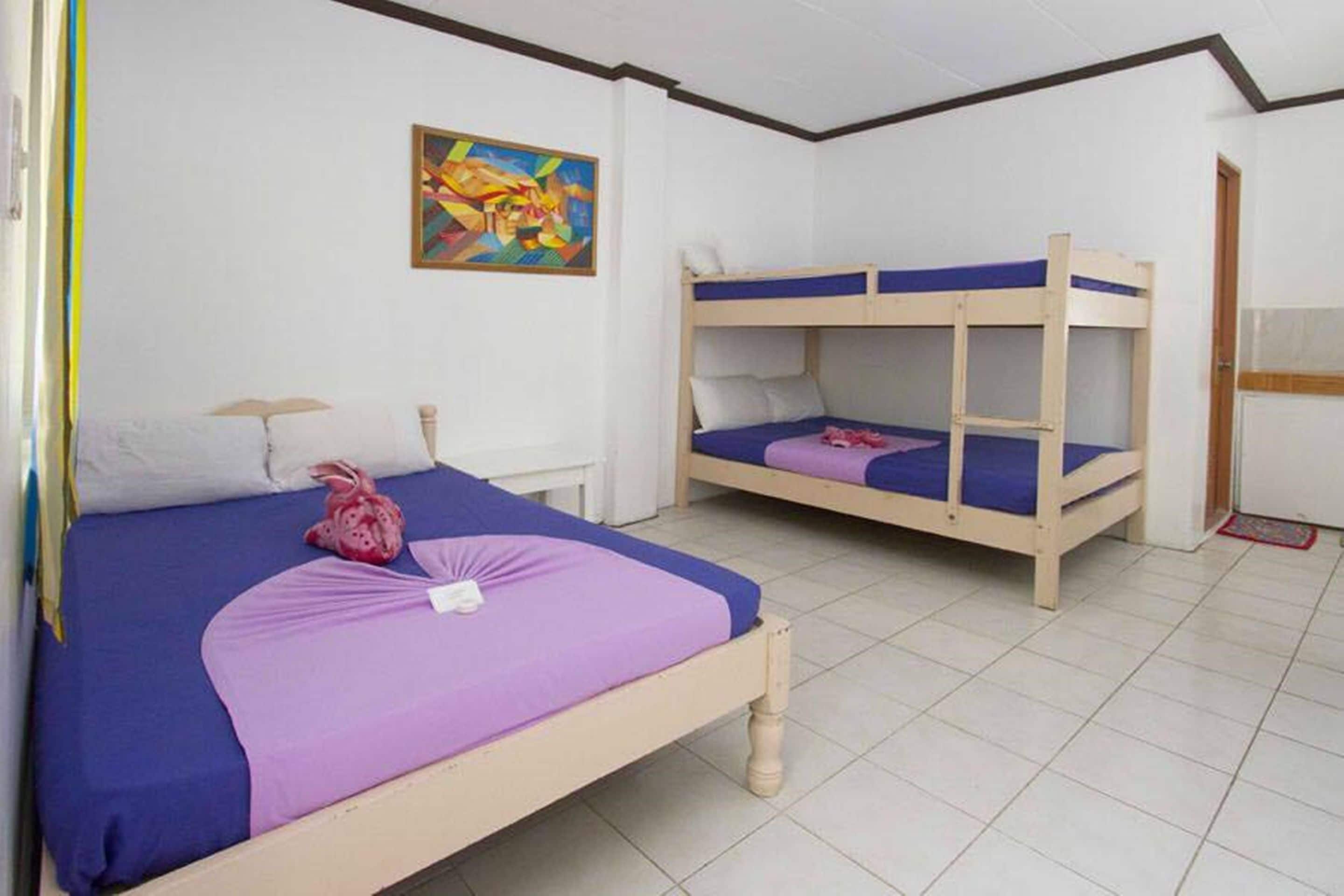A room with two bunk beds, a bed with a purple bedspread, a small table with a lamp, and a colorful abstract painting on the wall.