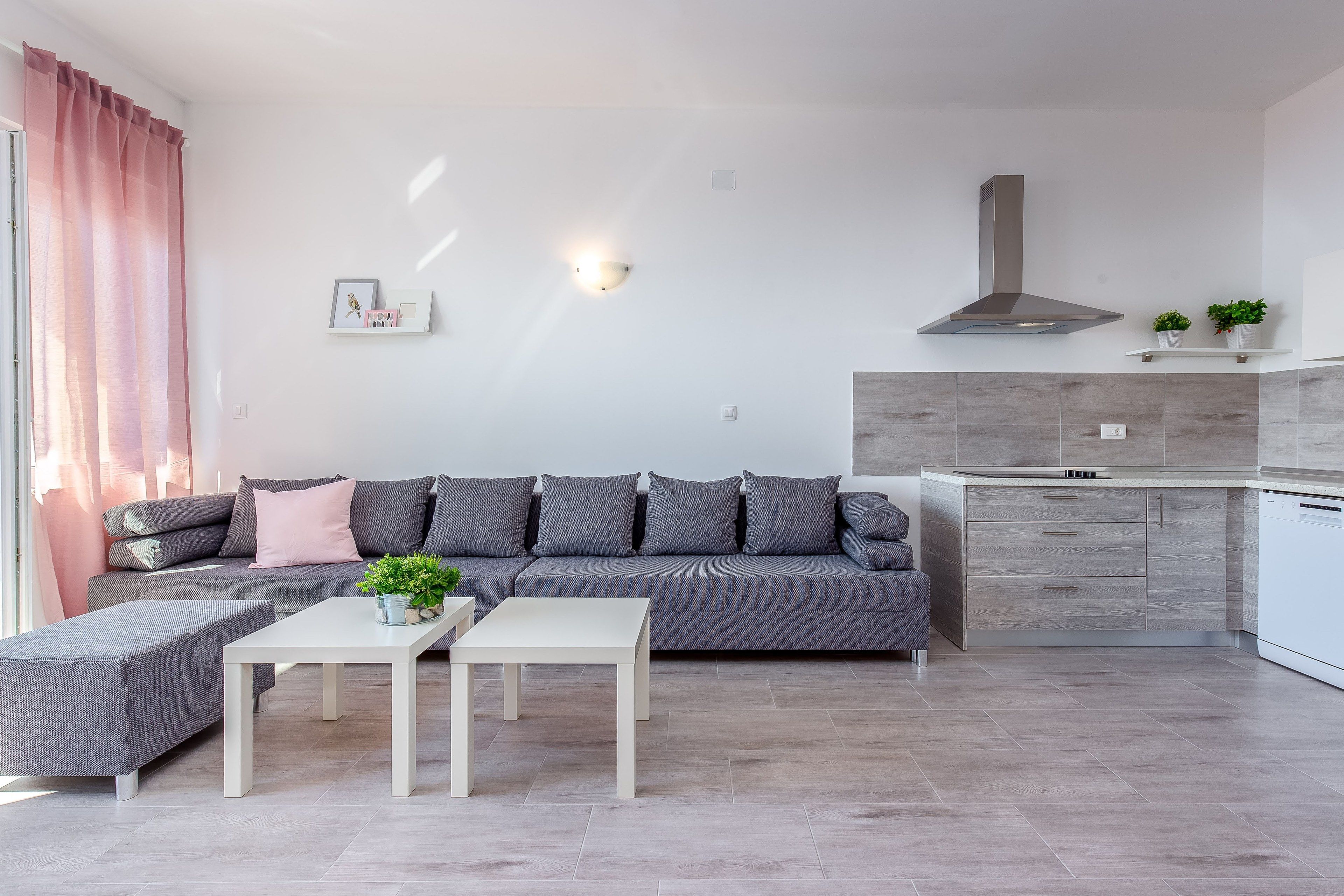 A modern living room with a grey sofa, a white coffee table, and a grey ottoman. There is a kitchen area with grey cabinets and a stainless steel range hood.