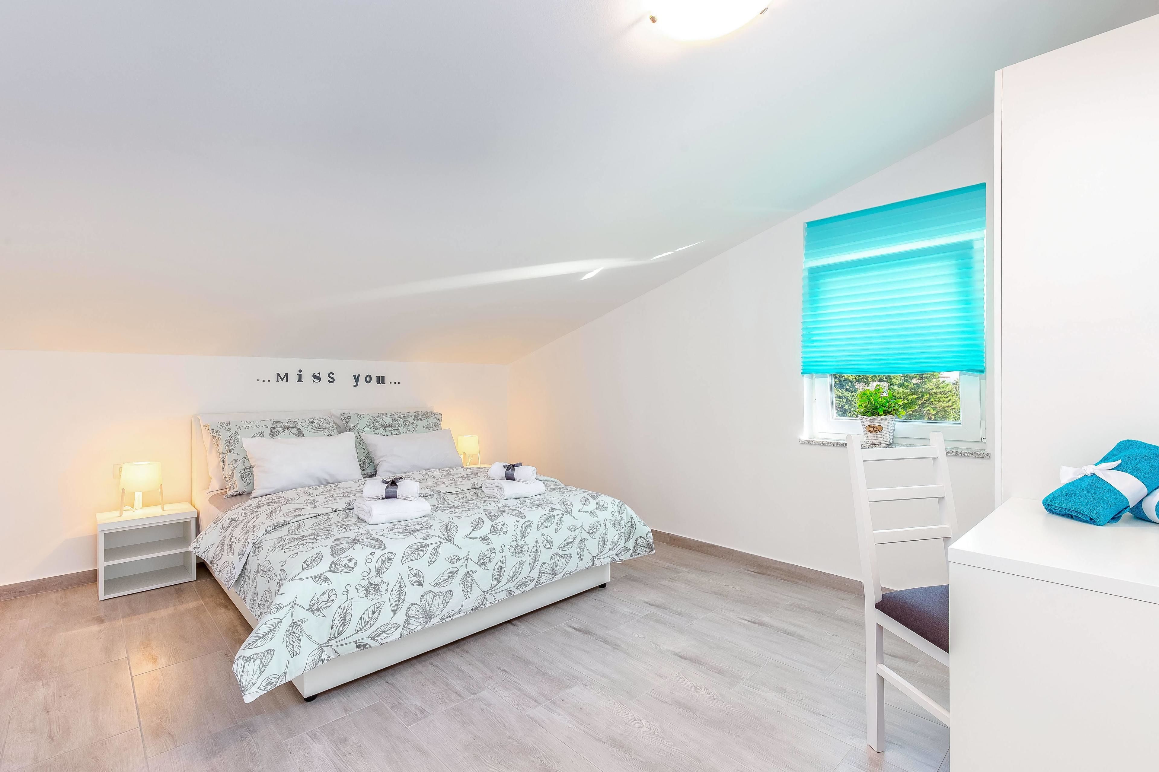 A bedroom with a bed, bedside tables, a window with blinds, and a view of greenery outside.