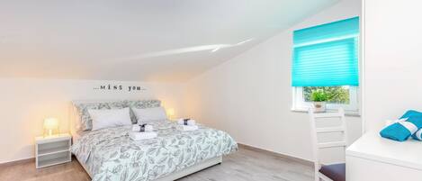 A bedroom with a bed, bedside tables, a window with blinds, and a view of greenery outside.