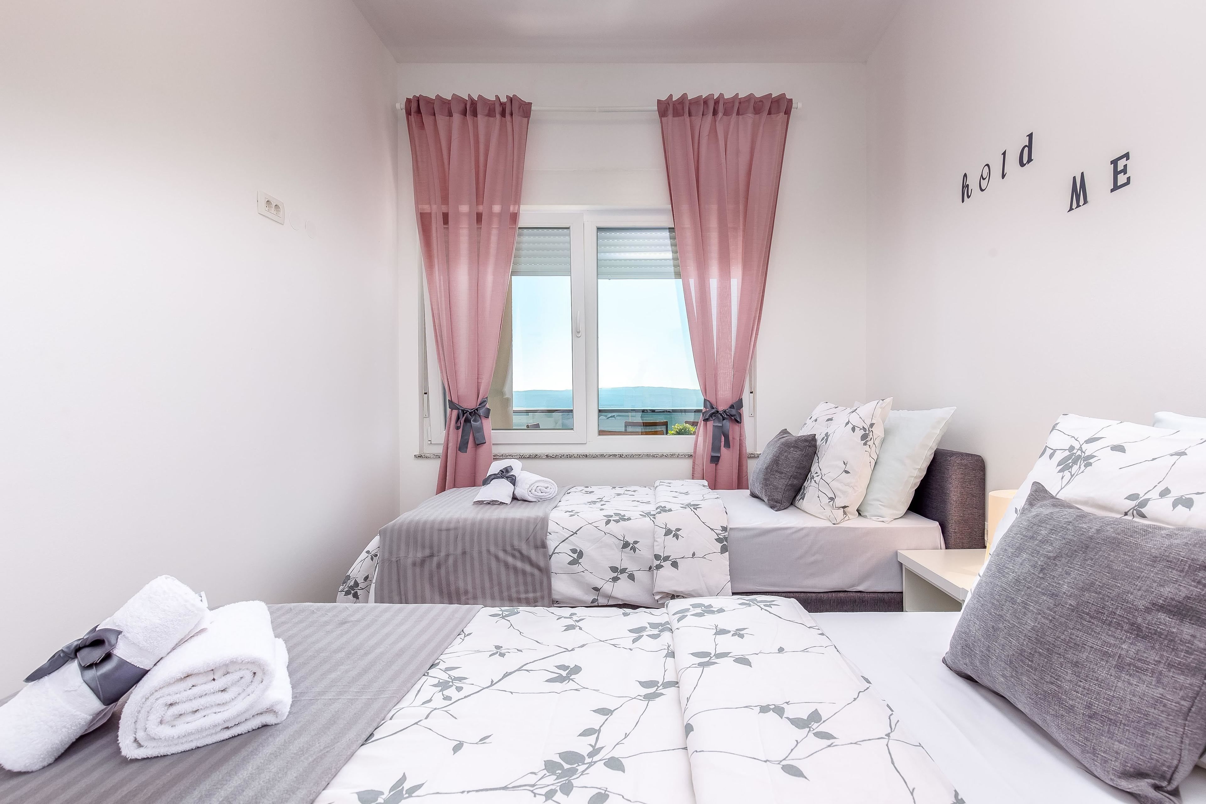 Un dormitorio con dos camas, cortinas rosas, vistas al exterior y una pared con el texto "hold ME".