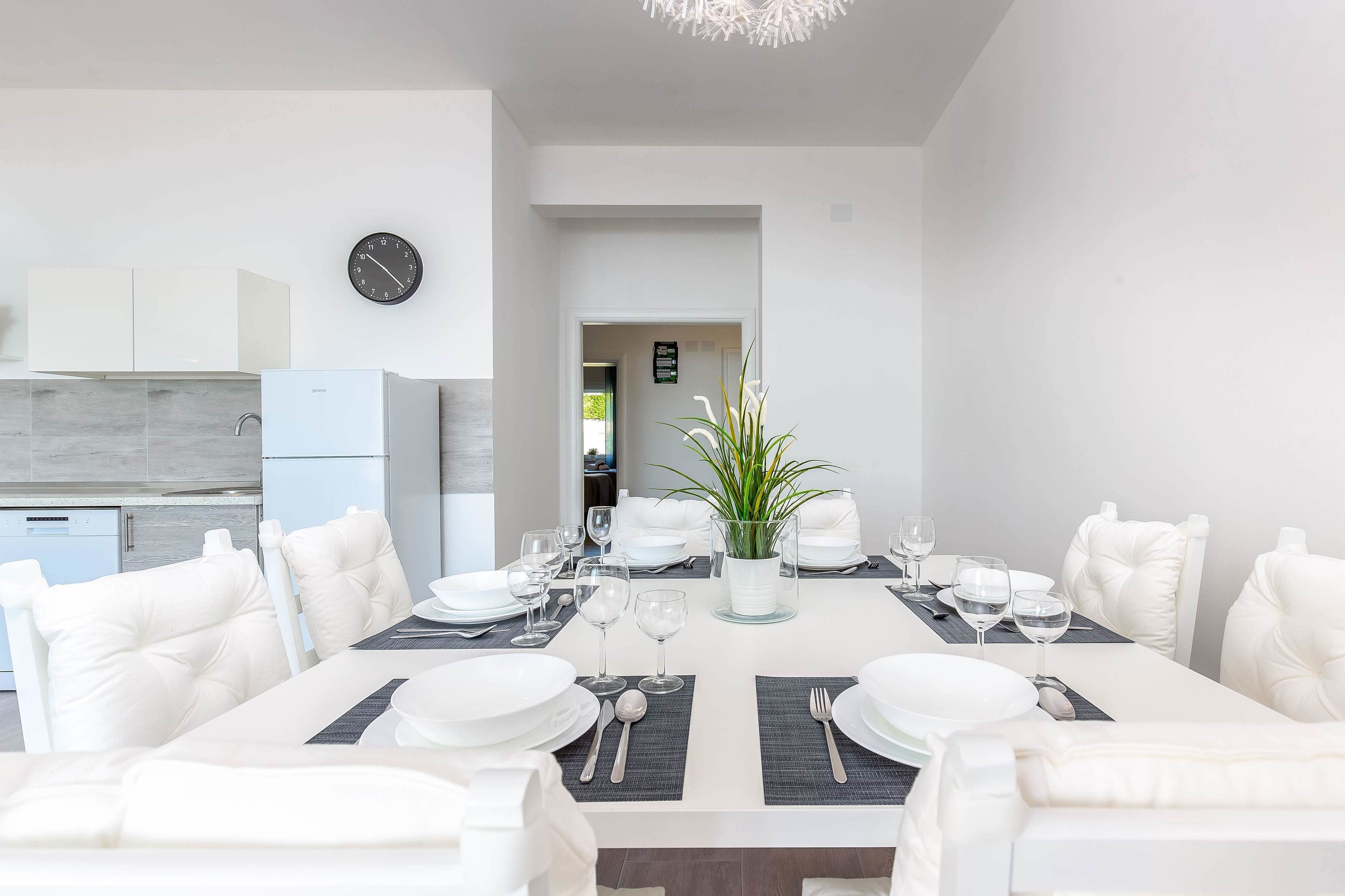 Un comedor moderno con mobiliario blanco, una mesa central dispuesta para seis personas y una cocina al fondo.