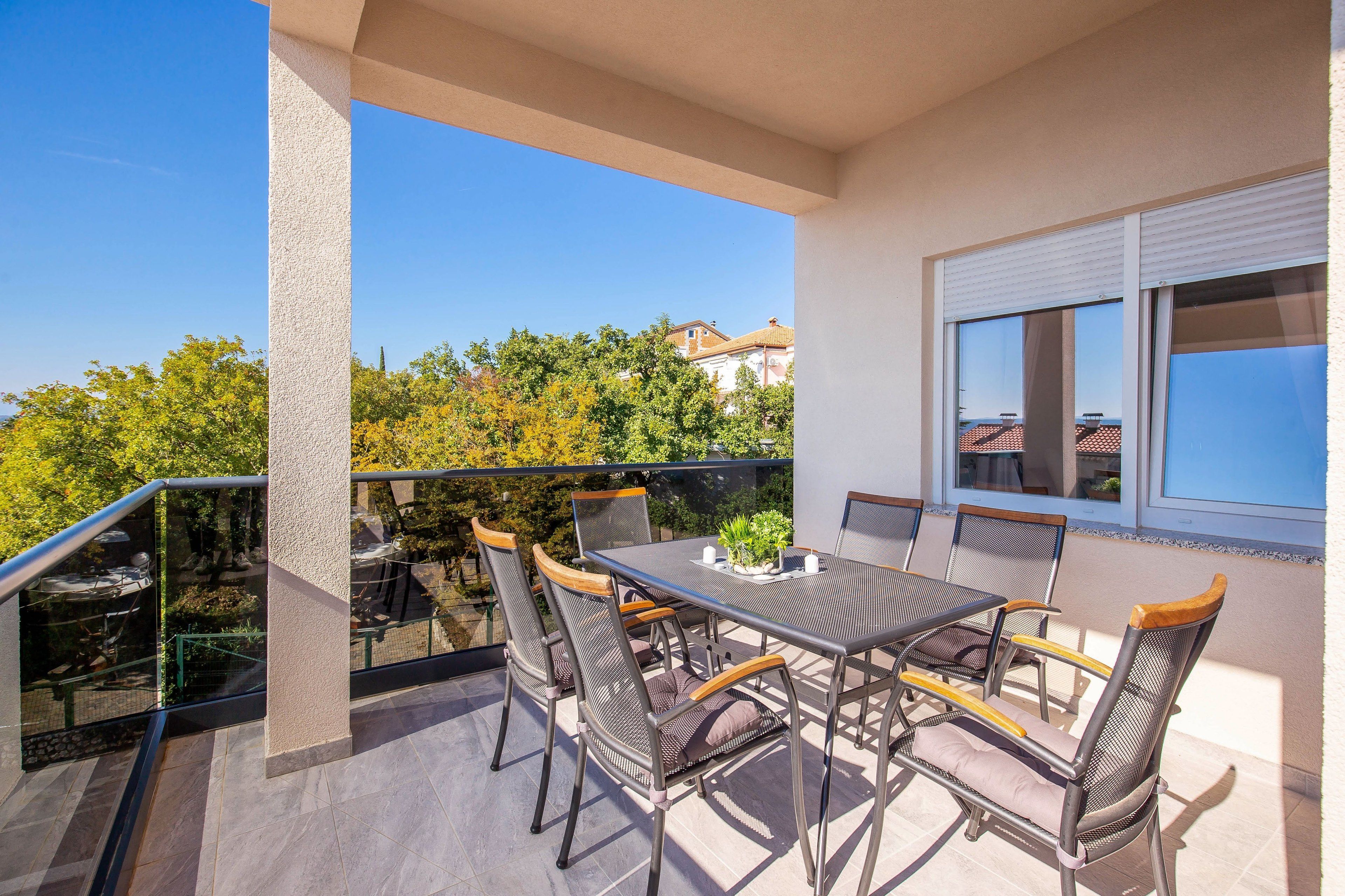 A balcony with a table and chairs, a glass railing, and a view of trees and buildings.