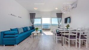 A modern living room with a blue sofa, dining table, and chairs, a flat-screen TV, and a large window with a view.