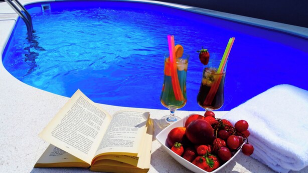 A poolside setting with a book, two drinks with straws, and a bowl of fruit.