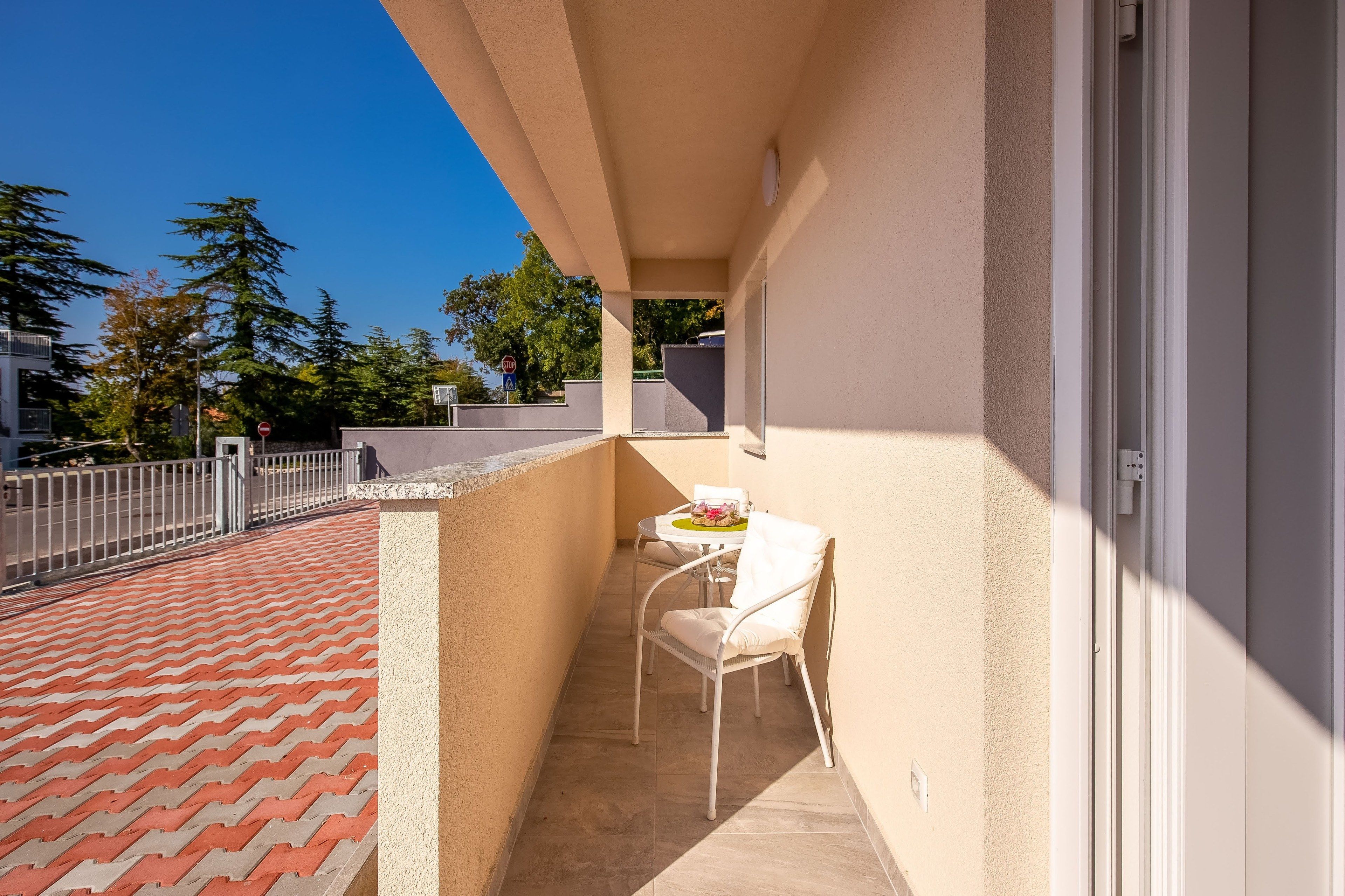 A balcony with a white table and chair, a red and beige tiled floor, and a view of trees and buildings.