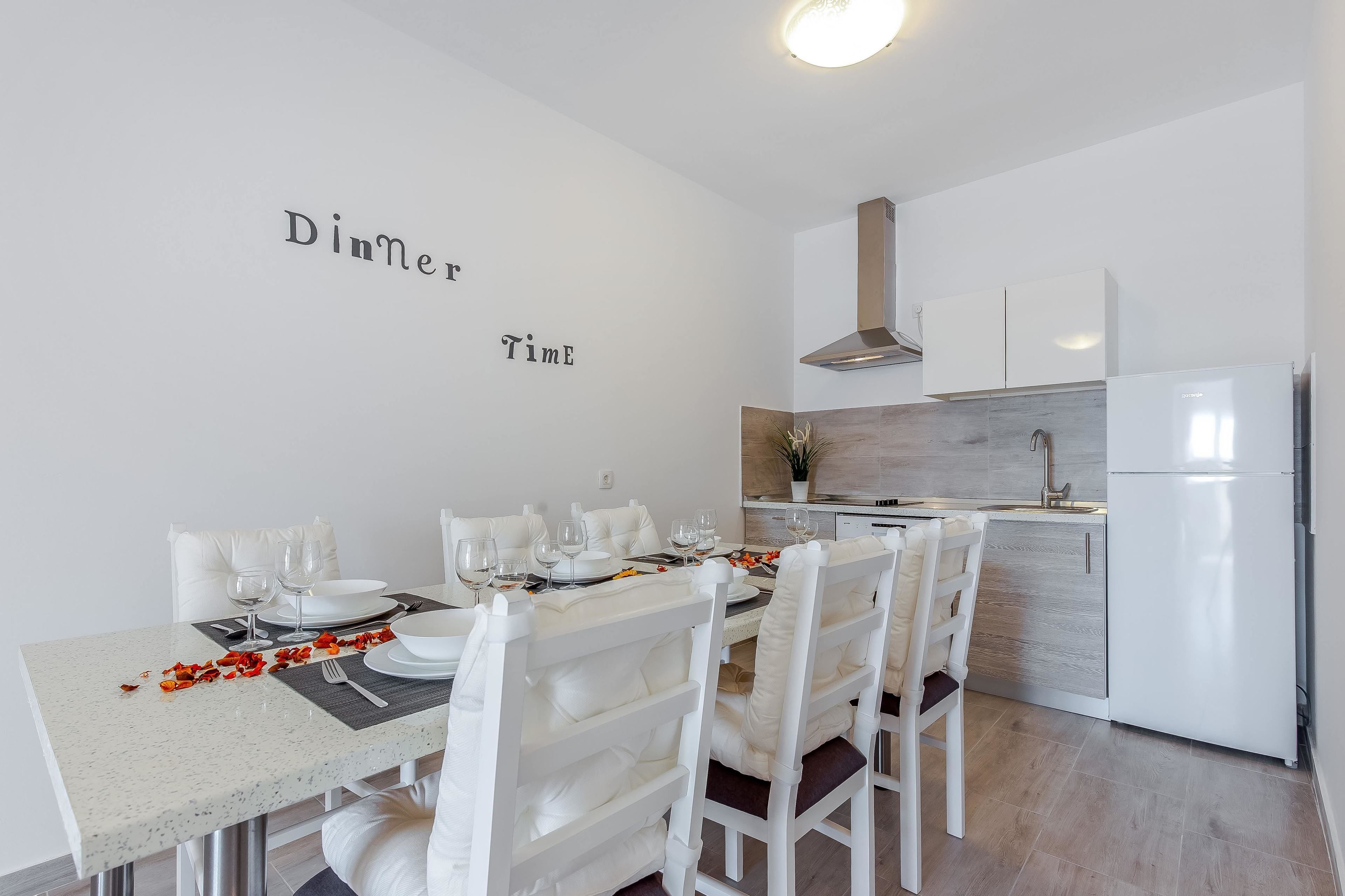 Un'area pranzo moderna con un tavolo bianco apparecchiato per due, sedie bianche e una cucina sullo sfondo.
