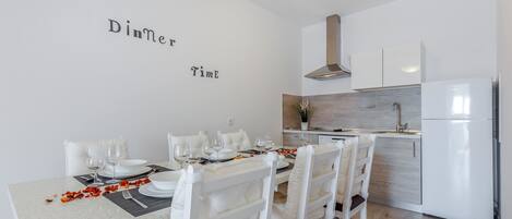 Un'area pranzo moderna con un tavolo bianco apparecchiato per due, sedie bianche e una cucina sullo sfondo.