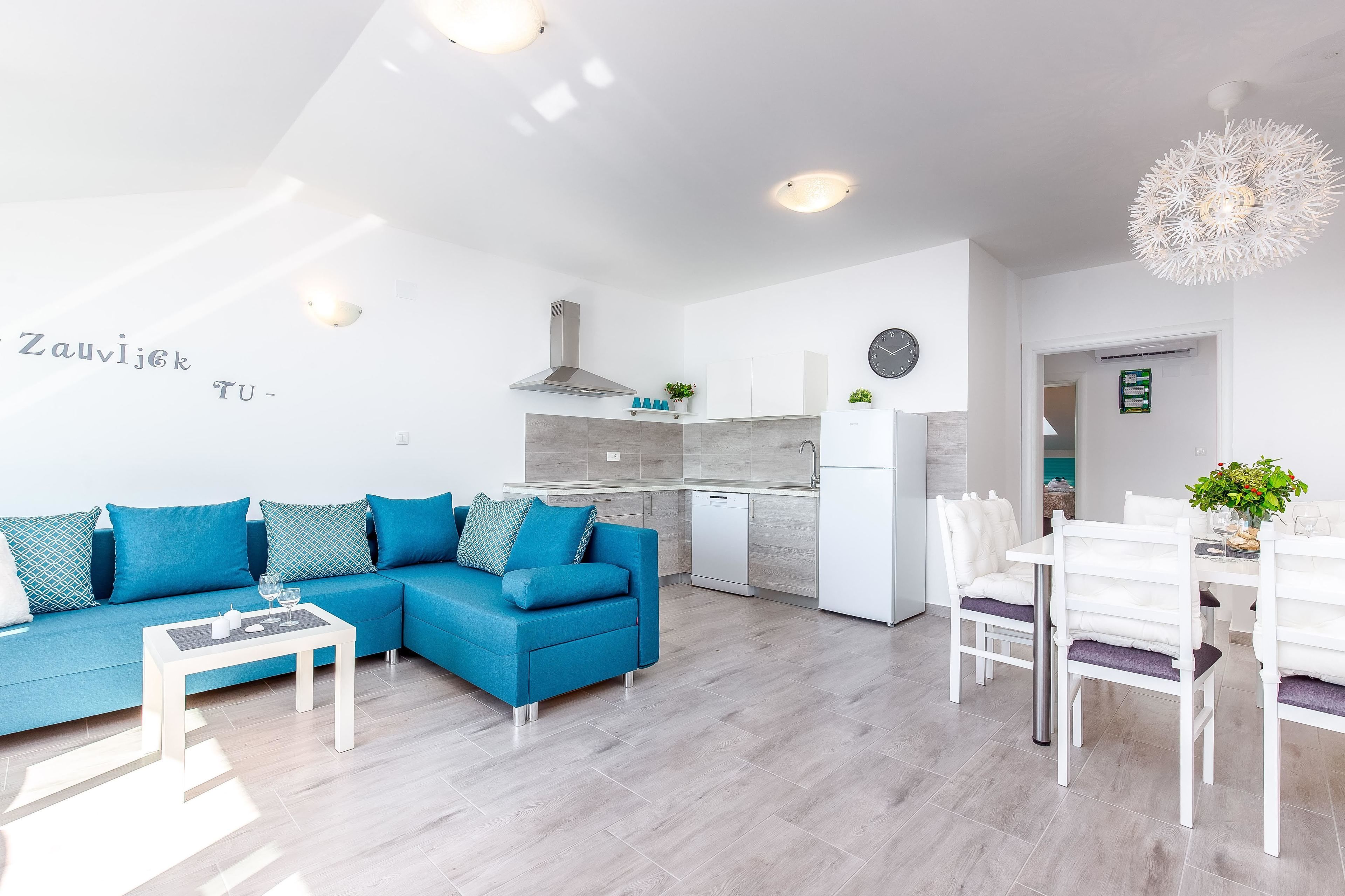 A modern living room with a blue sofa, a dining area with white chairs, and a kitchen with white cabinets.