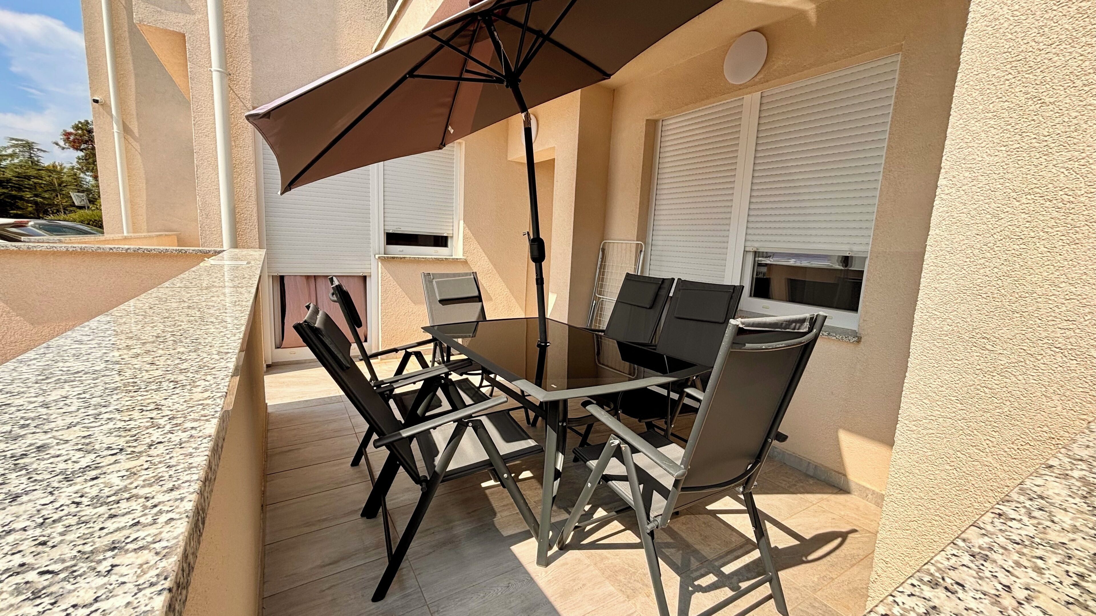 Terrassa exterior amb taula i caires de vidre, un parasol marró i una barbacoa.