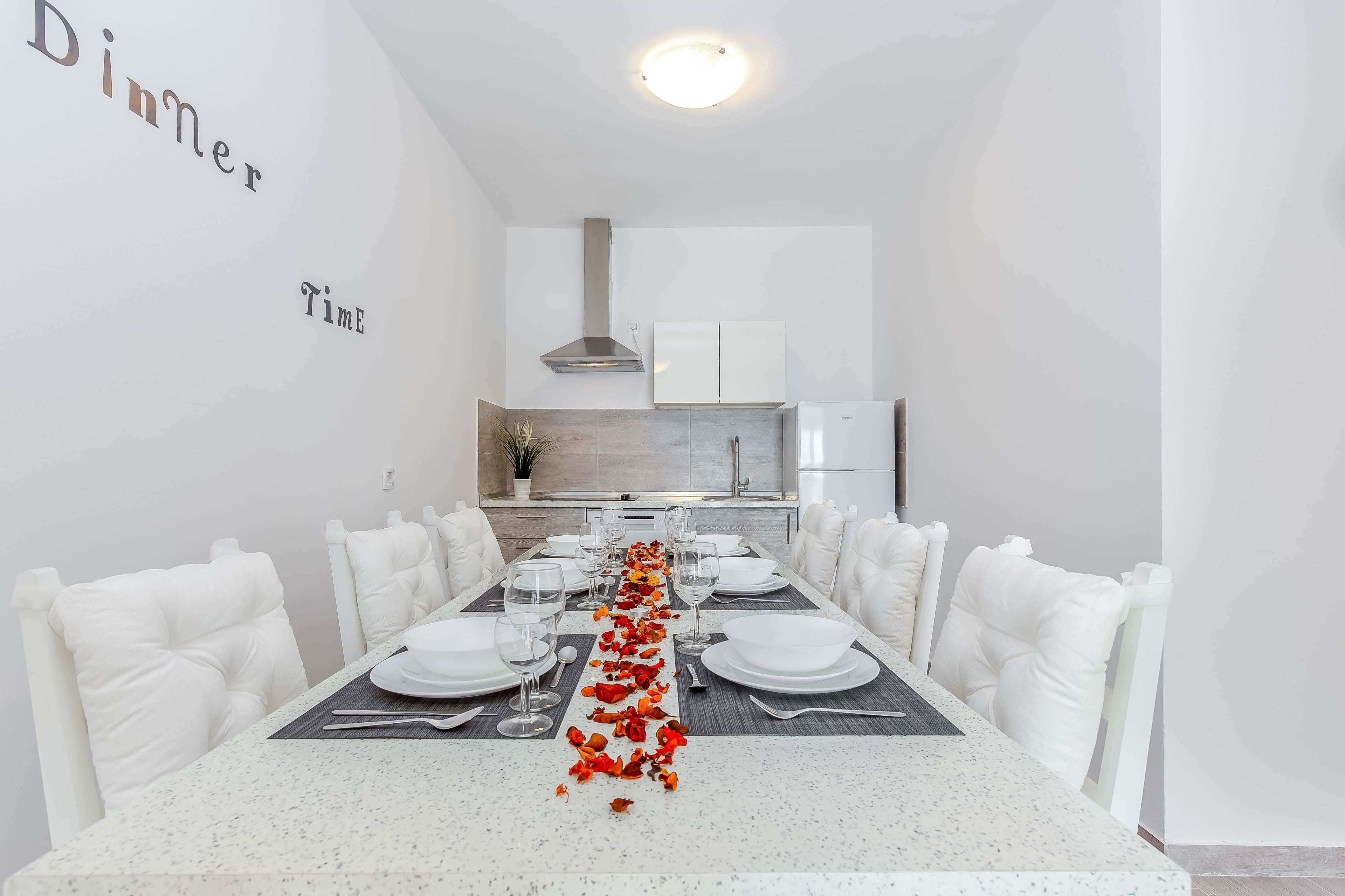A dining area with a long table set for dinner, white chairs, and a kitchen in the background.