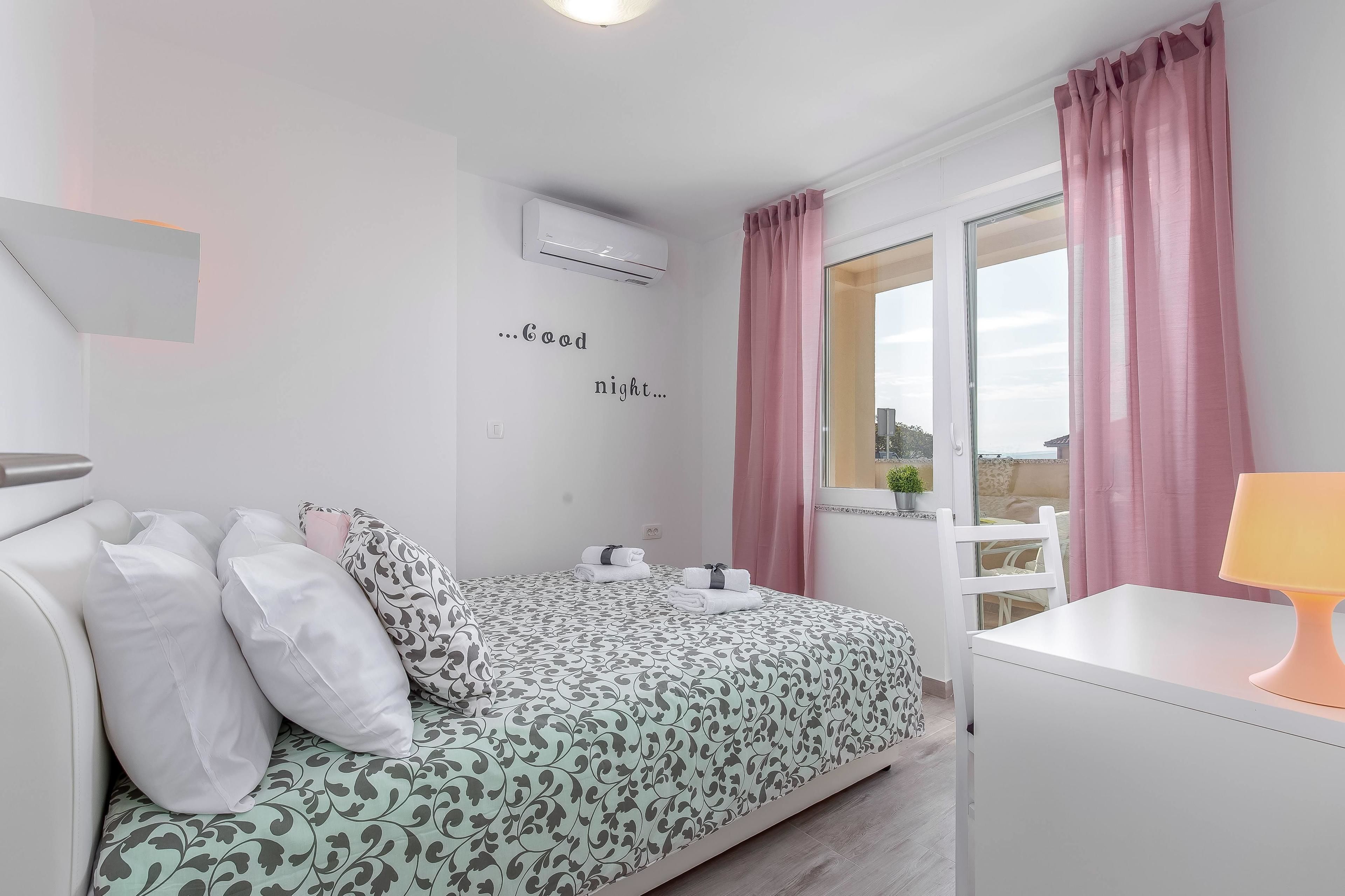 A bedroom with a bed, pillows, a bedside table, a lamp, a window with curtains, and a wall with a message.