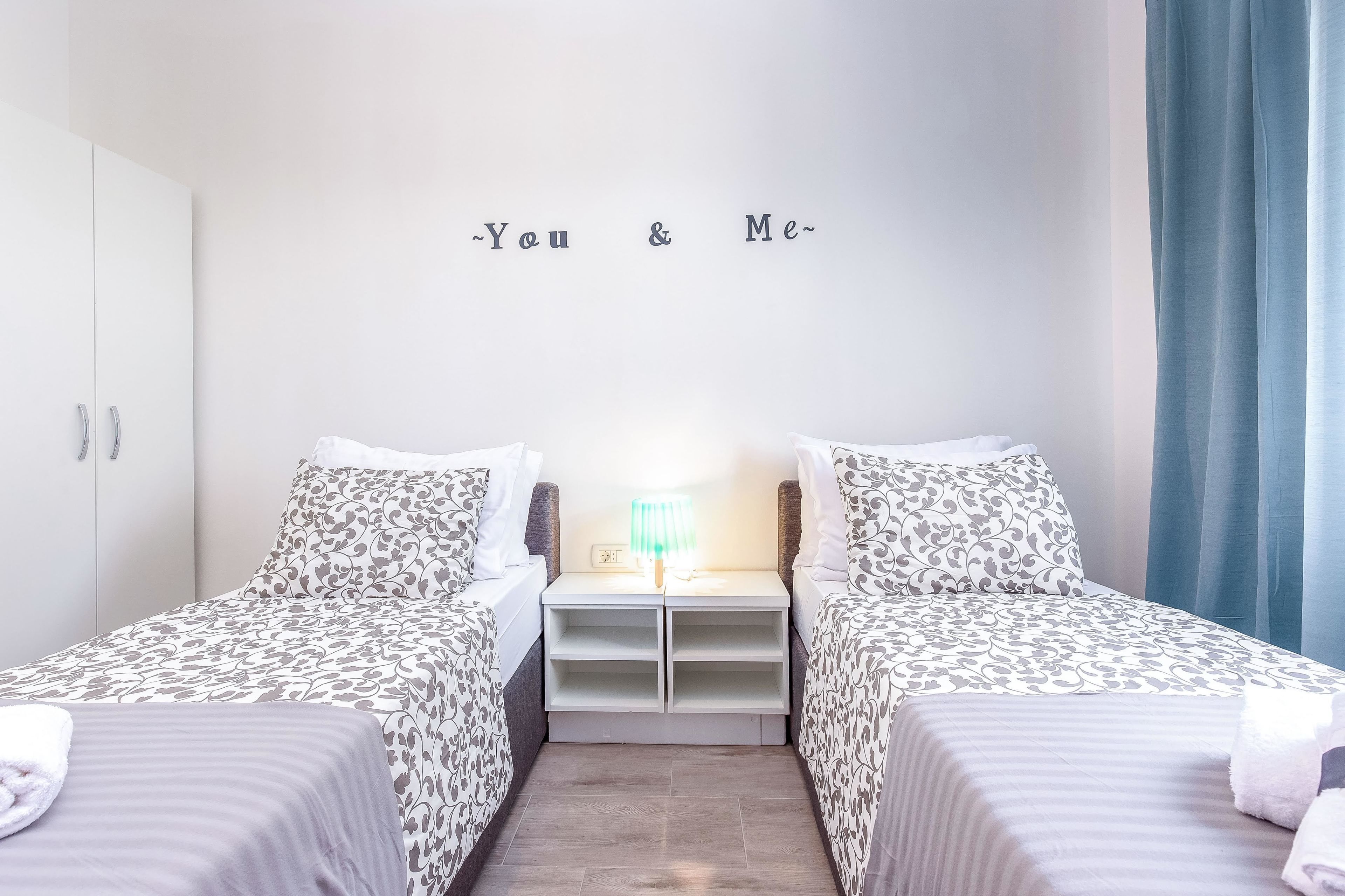 A twin room with two beds, a nightstand, a lamp, and a wall with the text "You & Me".