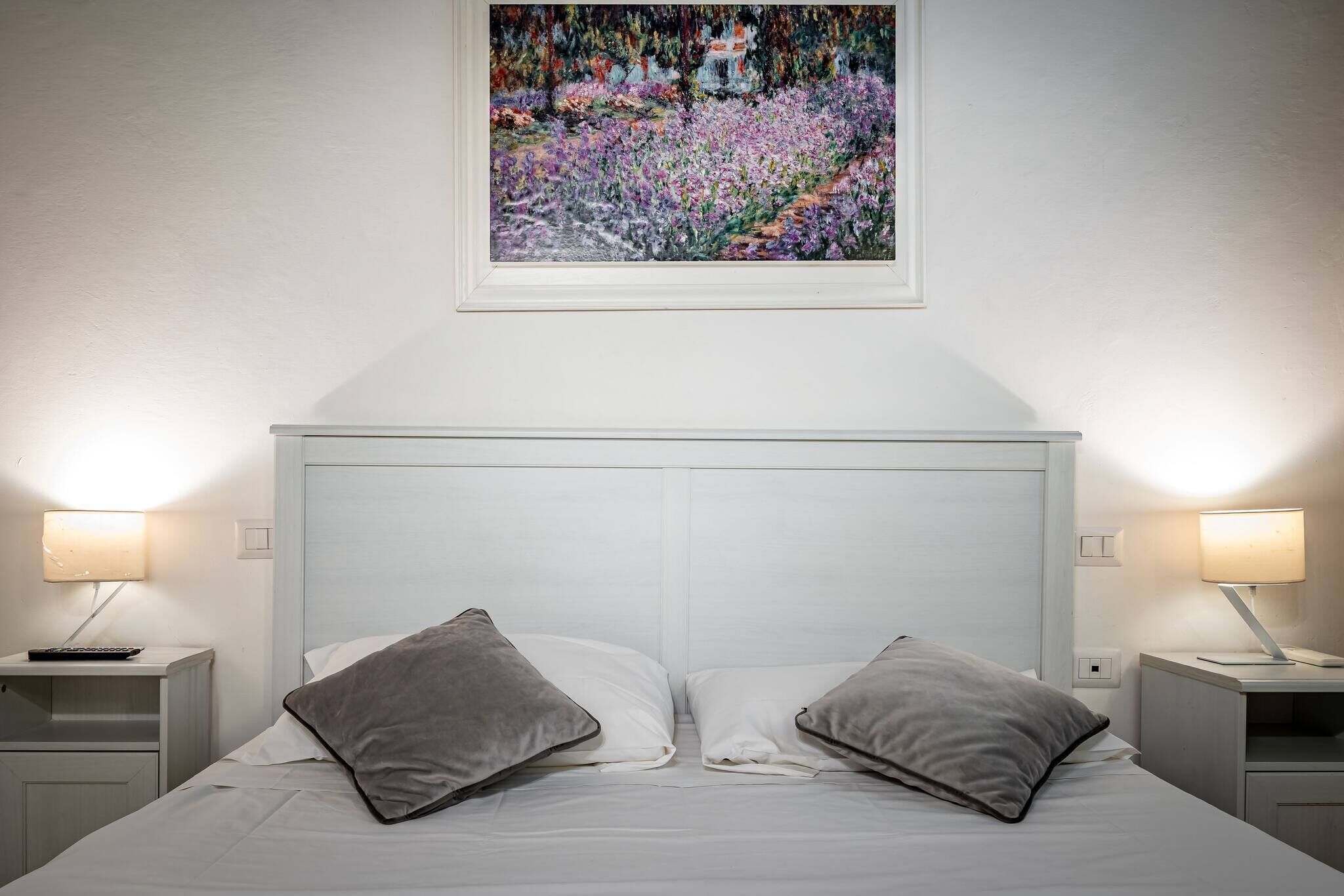 Une chambre à coucher avec un lit blanc, deux oreillers gris, une table de chevet avec une lampe et un tableau au-dessus du lit.