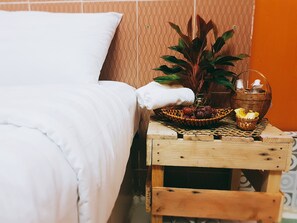 Una cama bien tendida con ropa de cama blanca, una mesita de noche de madera con una cesta tejida que contiene frutas y una botella, y una planta en maceta.