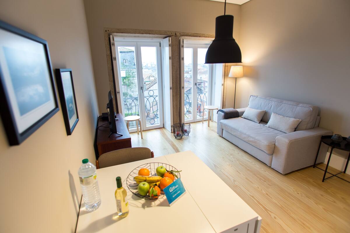 A modern living room with a sofa, a dining table with fruit and a water bottle, and a view through a glass door.