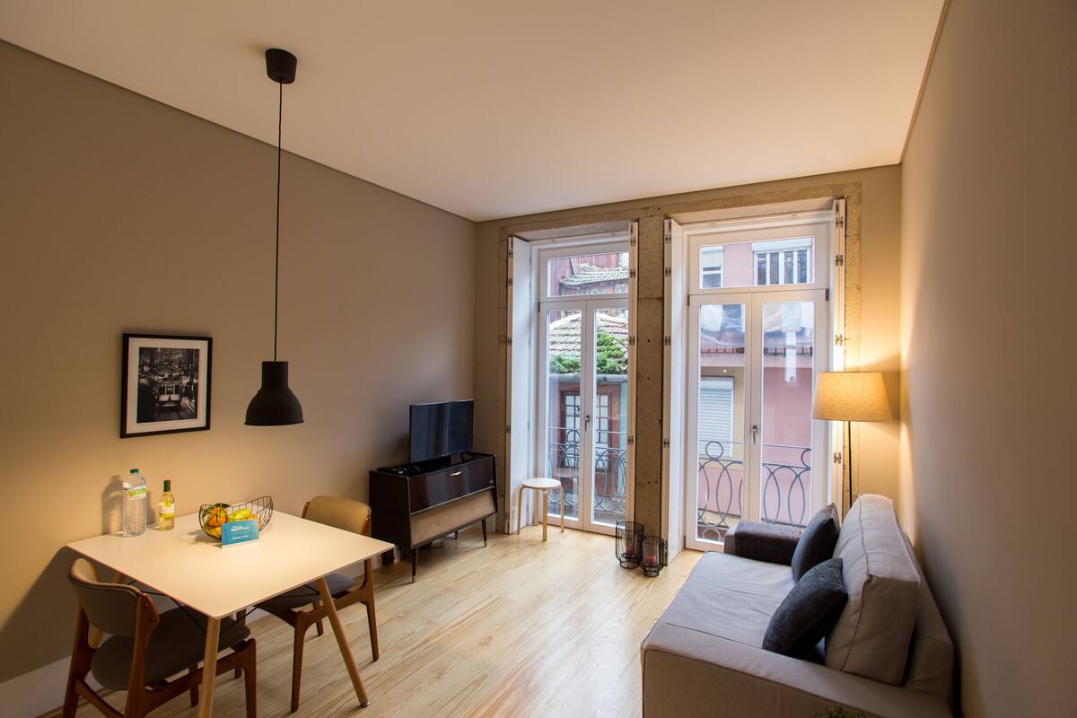 A compact living space with a dining table, chairs, a sofa, a TV, and a balcony with a view of buildings.