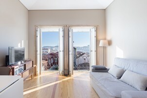 A modern living room with a sofa, TV, and a view through open doors to a balcony with a cityscape.