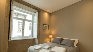 A bedroom with a bed, a nightstand, a lamp, a framed picture on the wall, and a window with a view of a building outside. - Porto and Clérigos Views by Porto City hosts (Porto)