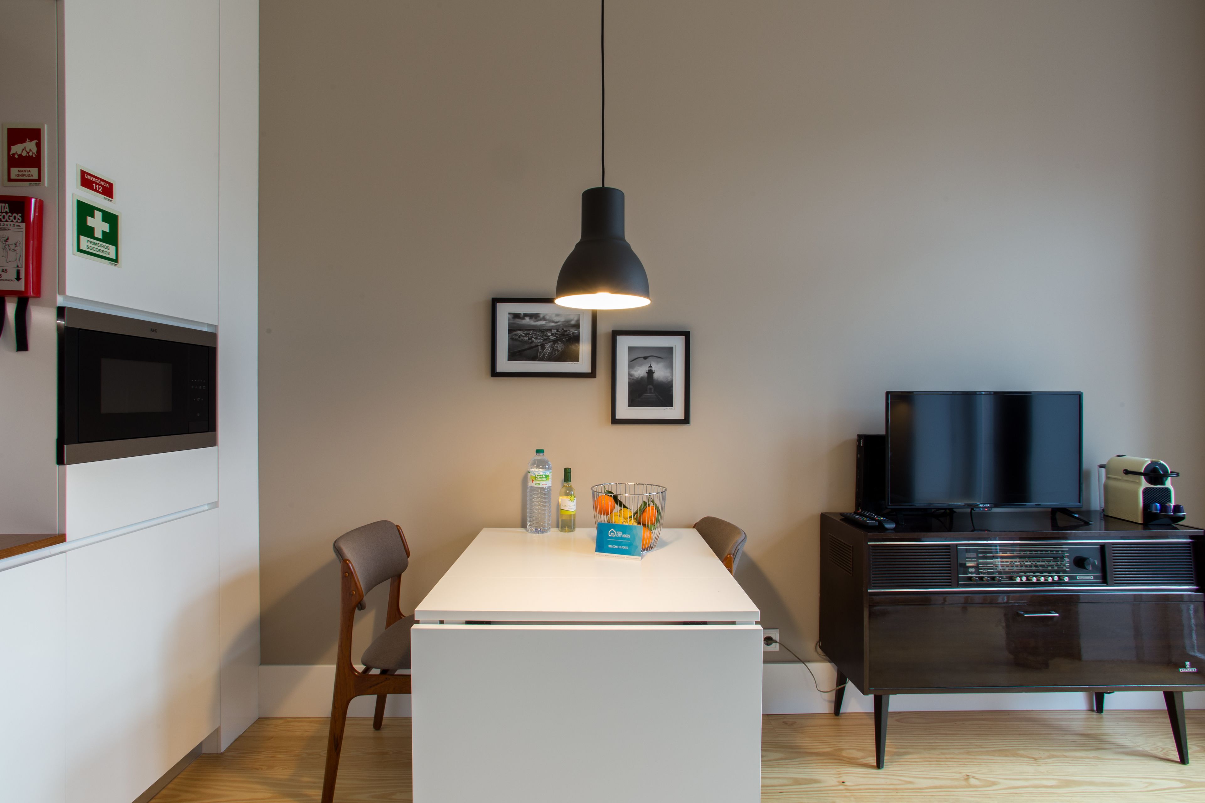 Eine moderne Küche mit einem weißen Kühlschrank, einer Mikrowelle und einem schwarzen Fernseher auf einem Holzschrank. Zwei Stühle, eine Pendelleuchte und gerahmte Bilder schmücken die Wand.