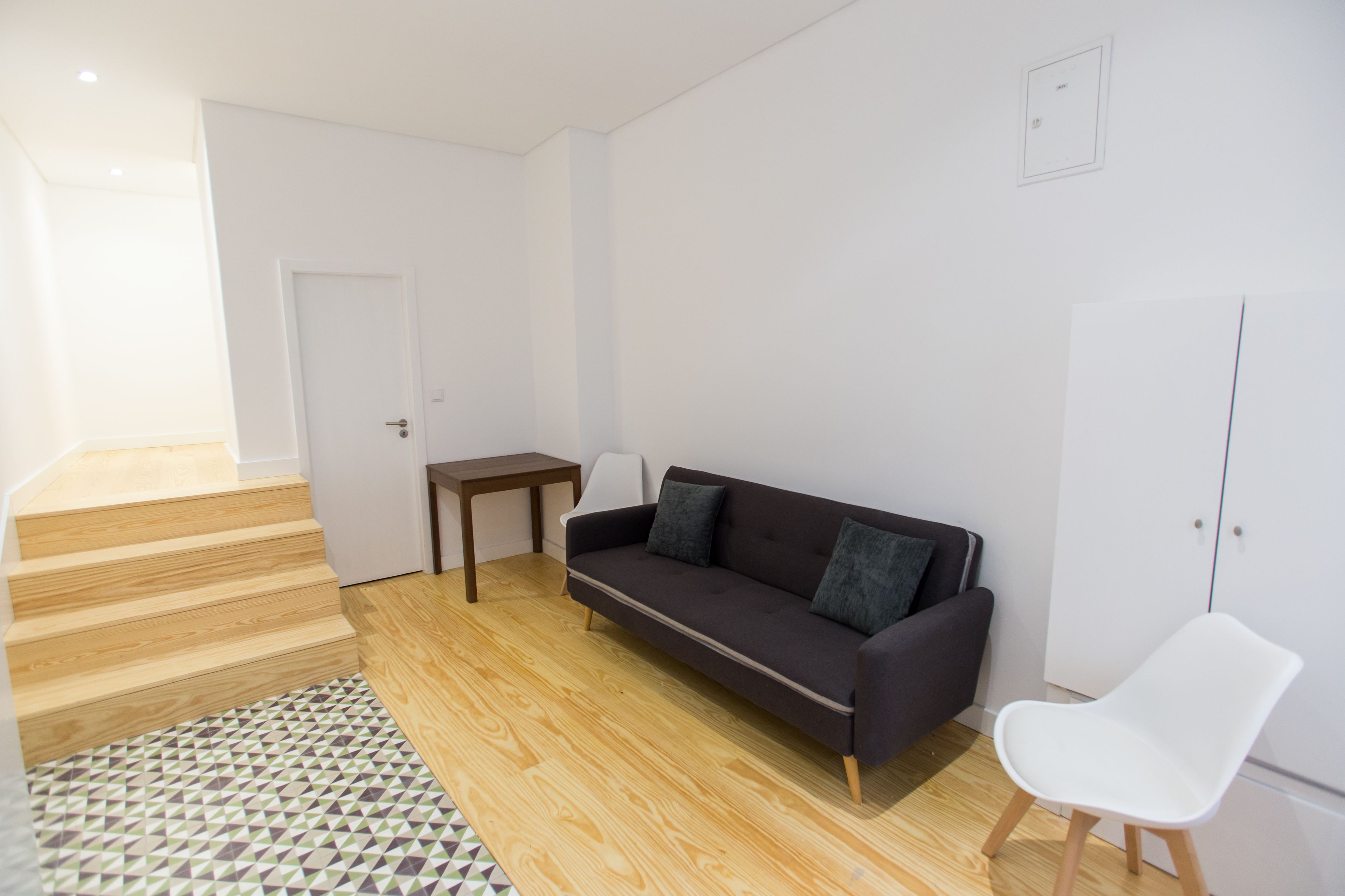 A modern living room with a sofa, a small table, a white chair, and a staircase leading to another room.