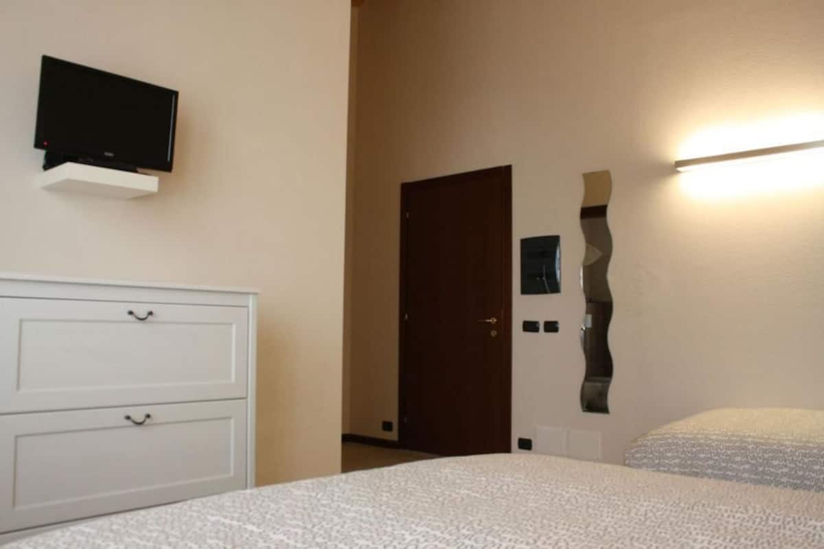 Een slaapkamer met een bed, een kastje, een televisie aan de muur en een deur.