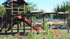 A wooden play structure with a slide and climbing elements, set against a backdrop of a clear sky and greenery. - Cascina Bosco Gerolo (Rivergaro)