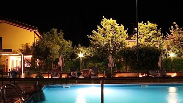 A swimming pool with lounge chairs and umbrellas, surrounded by trees and a building with lights. - Cascina Bosco Gerolo (Rivergaro)