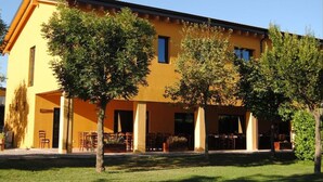 A yellow building with a covered outdoor seating area and trees in front. - Cascina Bosco Gerolo (Rivergaro)
