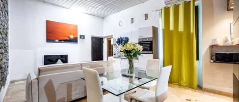 A modern living room with a glass dining table, white sofas, a flat-screen TV, and a kitchen area with a yellow curtain.