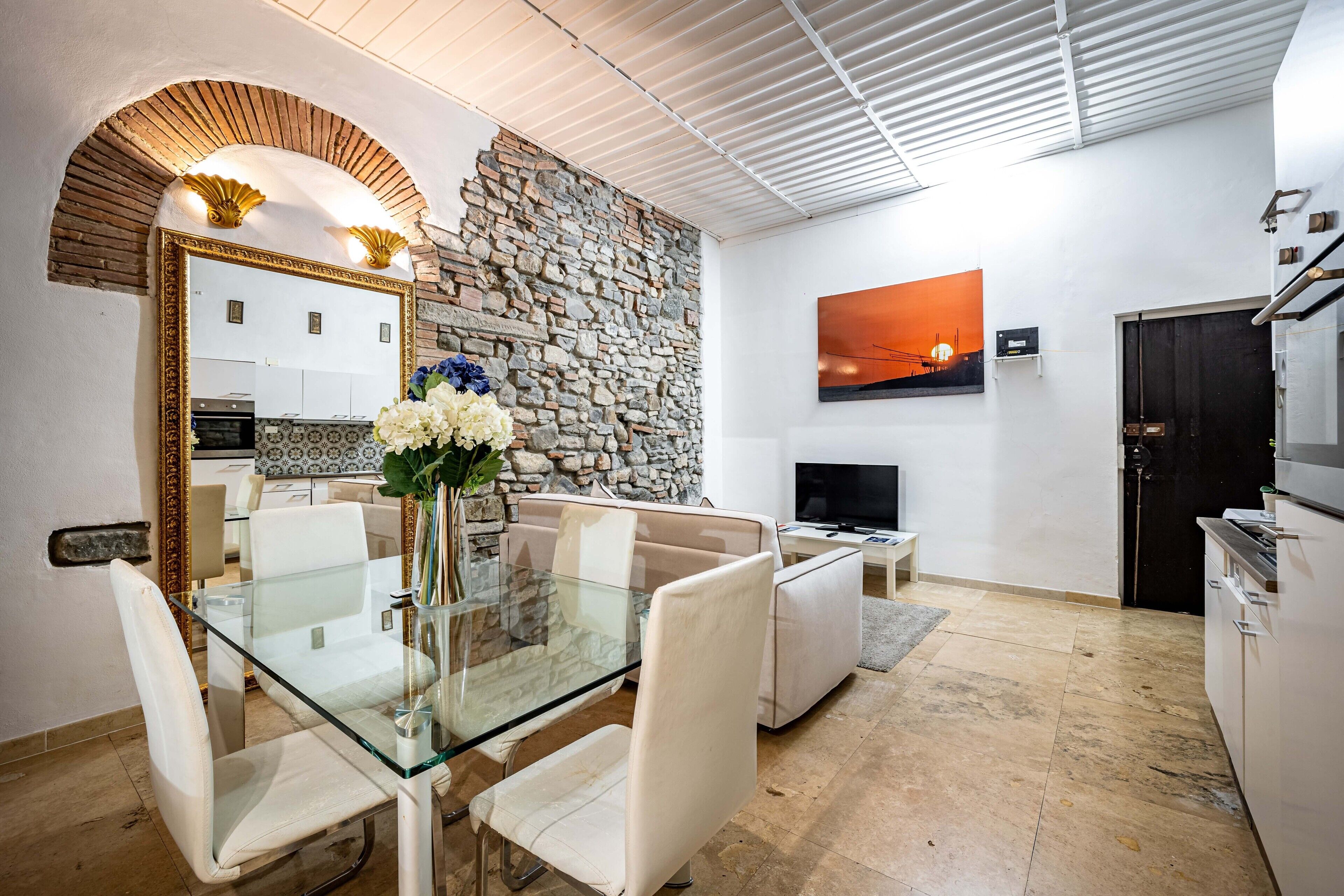 A modern living room with a glass dining table, white sofas, a stone wall, a TV, and a kitchen area.