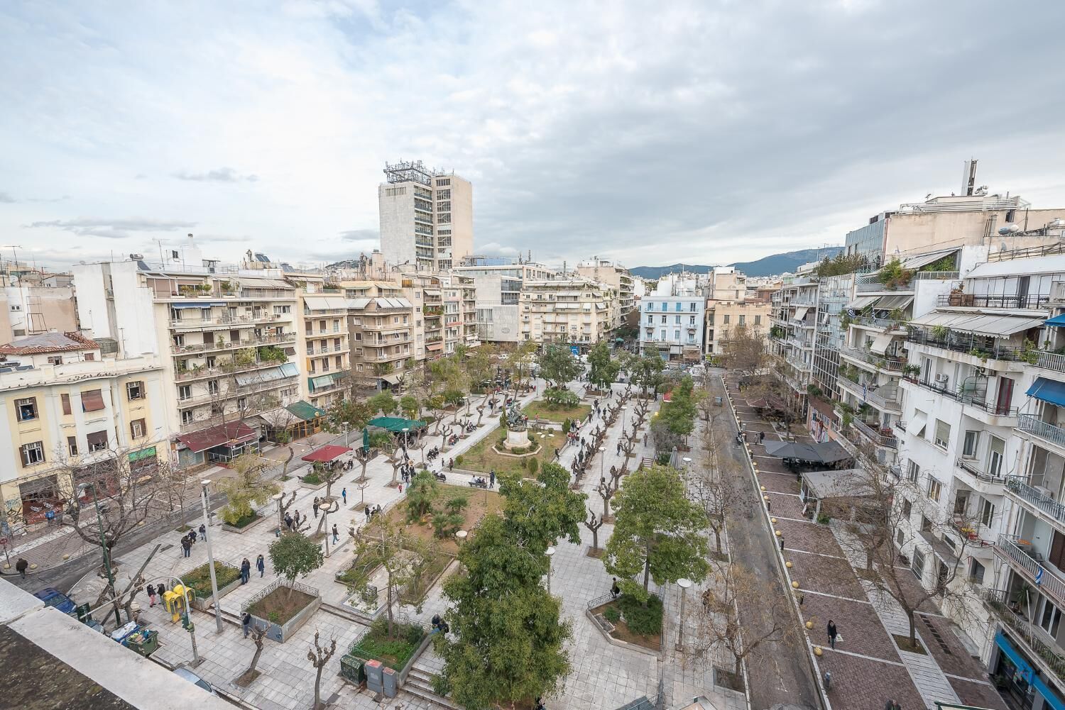 Une vue urbaine comprenant un mélange de bâtiments résidentiels, une place centrale et ouverte bordée d’arbres, et un grand immeuble en arrière-plan.