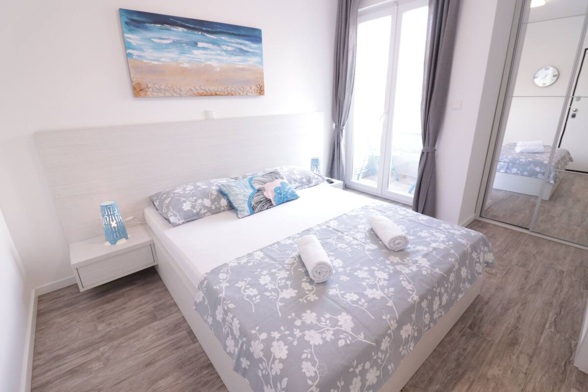 A bedroom with a bed, a nightstand, a painting, and a window with curtains.