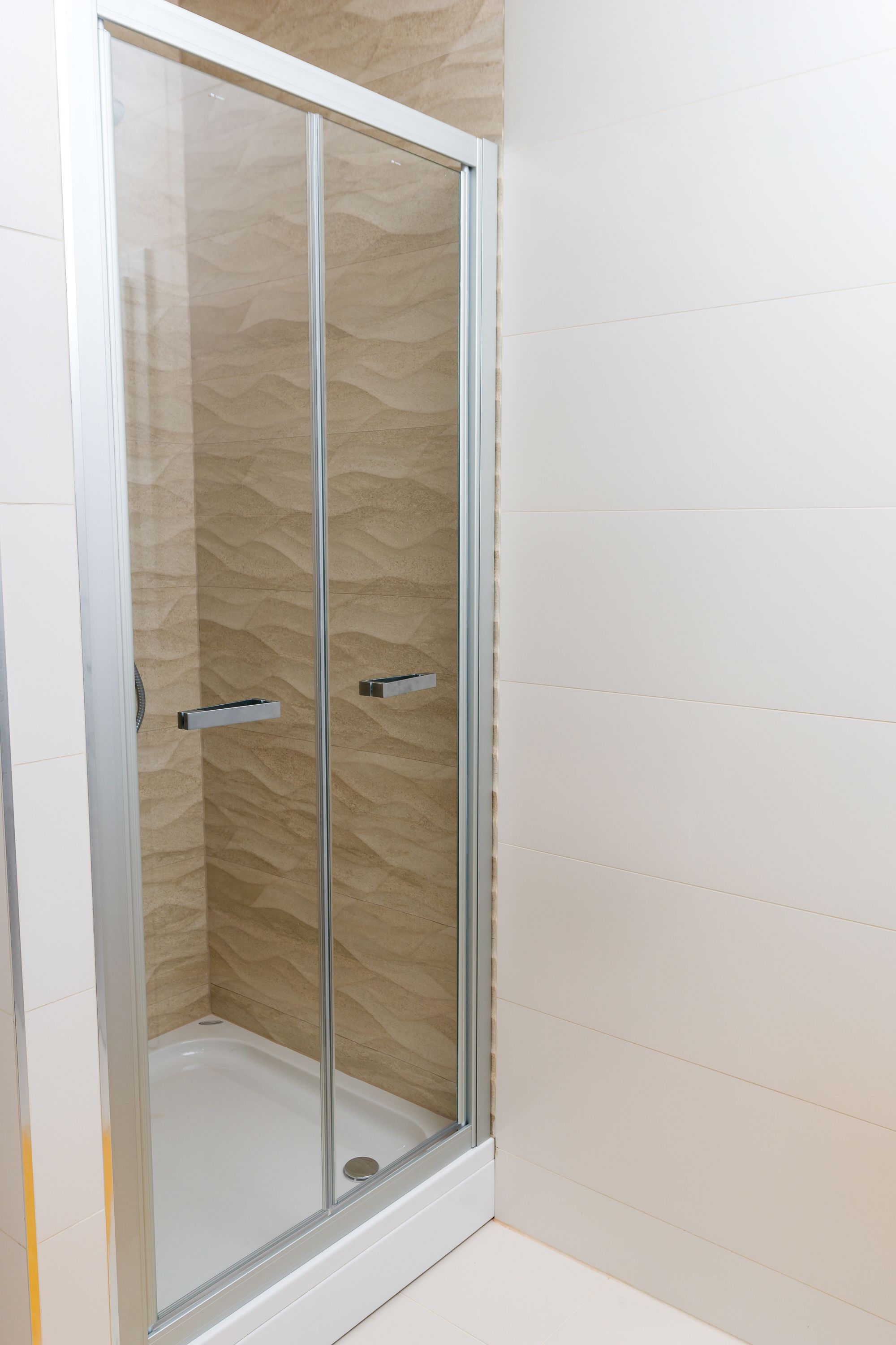 A glass-enclosed shower stall with a beige textured wall and a white floor.