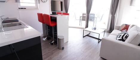 A modern apartment with a kitchen, dining area, and living room. It includes a white sofa, a dining table with red chairs, and a TV mounted on the wall.