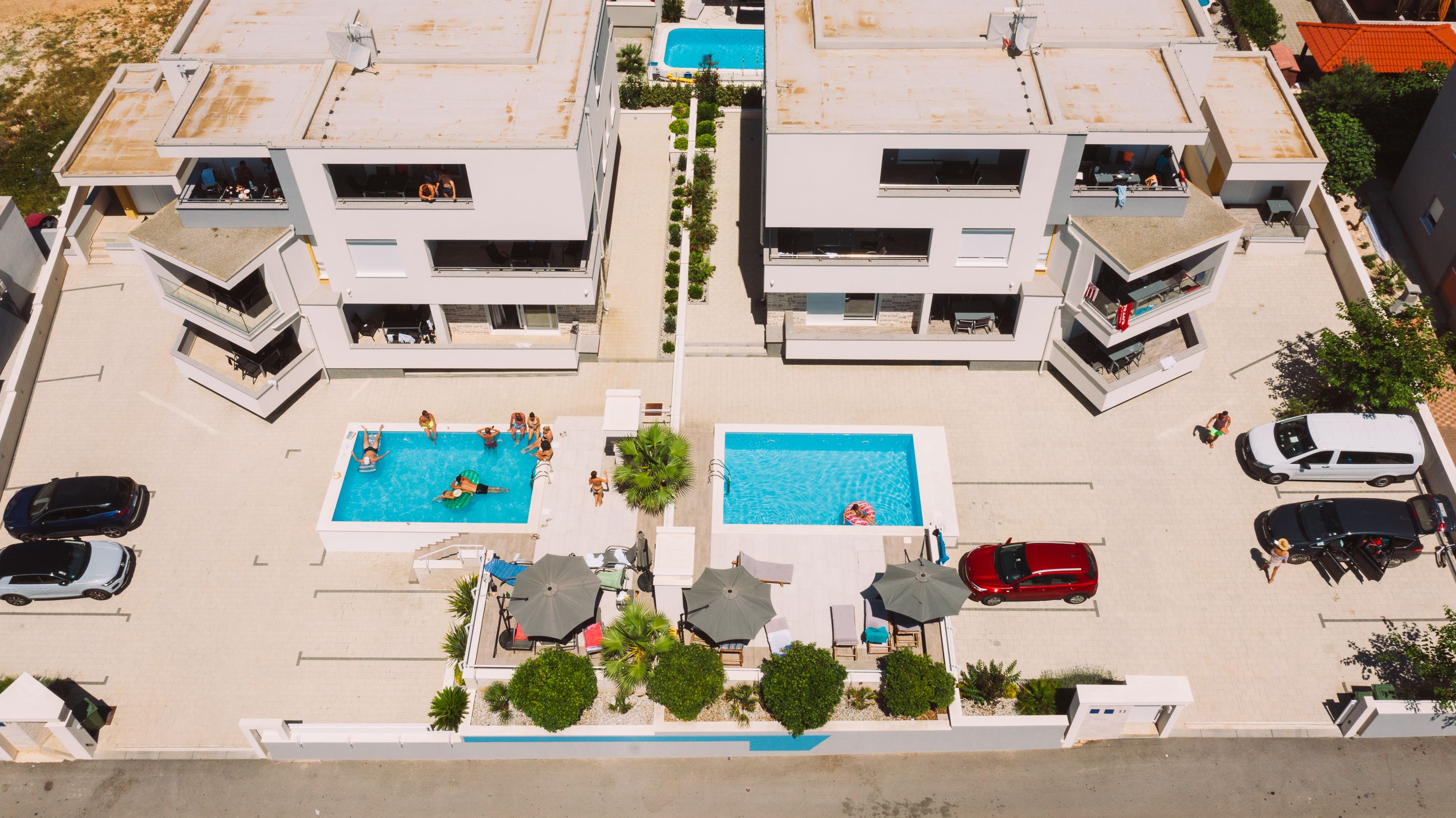 An aerial view of a modern building with two swimming pools, surrounded by parked cars and a few people.