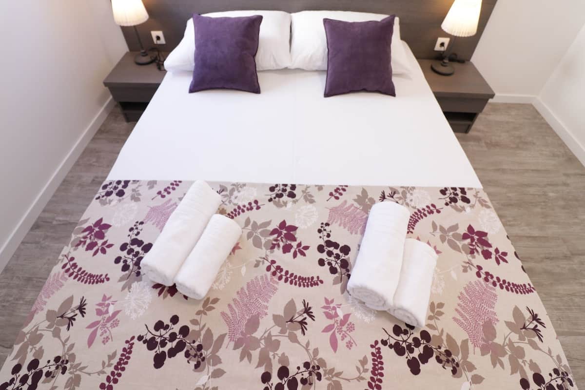 A neatly made bed with two purple pillows, a floral-patterned bedspread, and two rolled towels on a wooden floor.