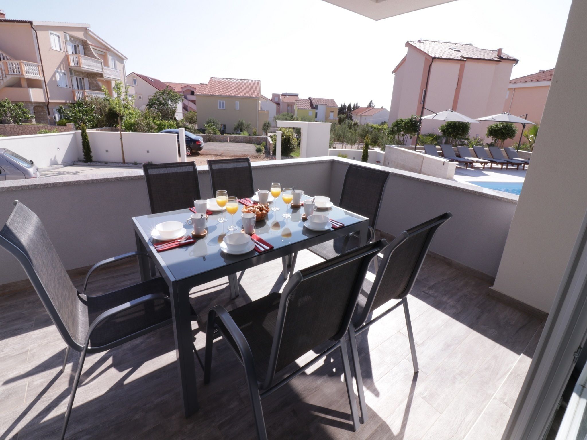 A dining table set for breakfast on a balcony with chairs, a view of neighboring buildings, and a pool in the background.