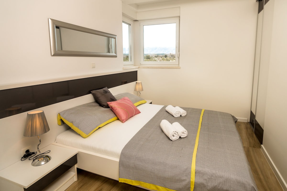 A modern hotel room with a bed, bedside lamps, a mirror, and a view of the city through the window.