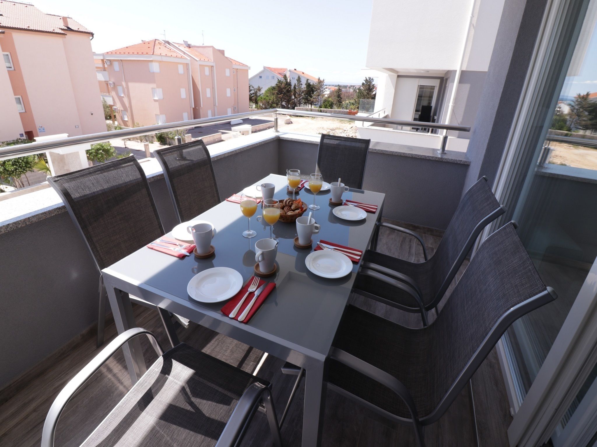 Una mesa de comedor con platos, cubiertos y vasos, rodeada de sillas, en un balcón con vistas a edificios.