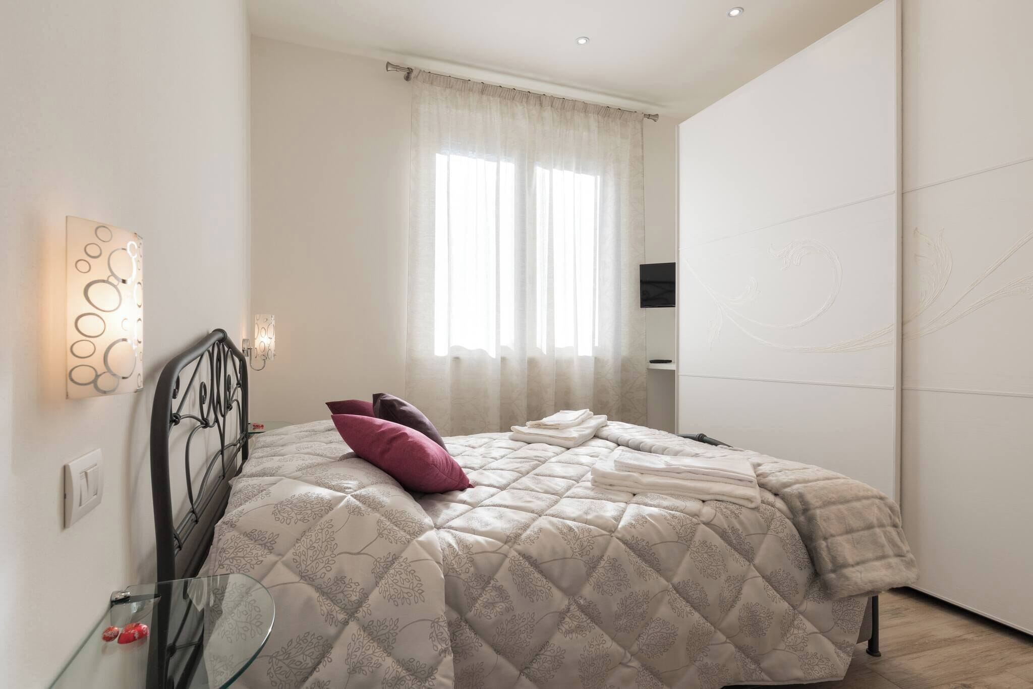 Un dormitorio con cama, una mesita de vidrio, televisión montada en la pared y ventana con cortinas.