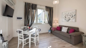 Una sala de estar con un sofá, una mesa de comedor con sillas, un televisor montado en la pared y una ventana con cortinas.