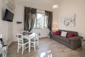 A living room with a dining table, white chairs, a grey sofa, a TV mounted on the wall, and a window with curtains.