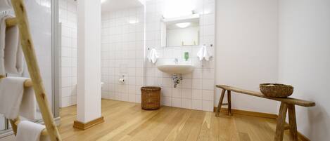Un baño con suelo de madera, azulejos blancos, un lavabo, un espejo y un banco de madera con una cesta.
