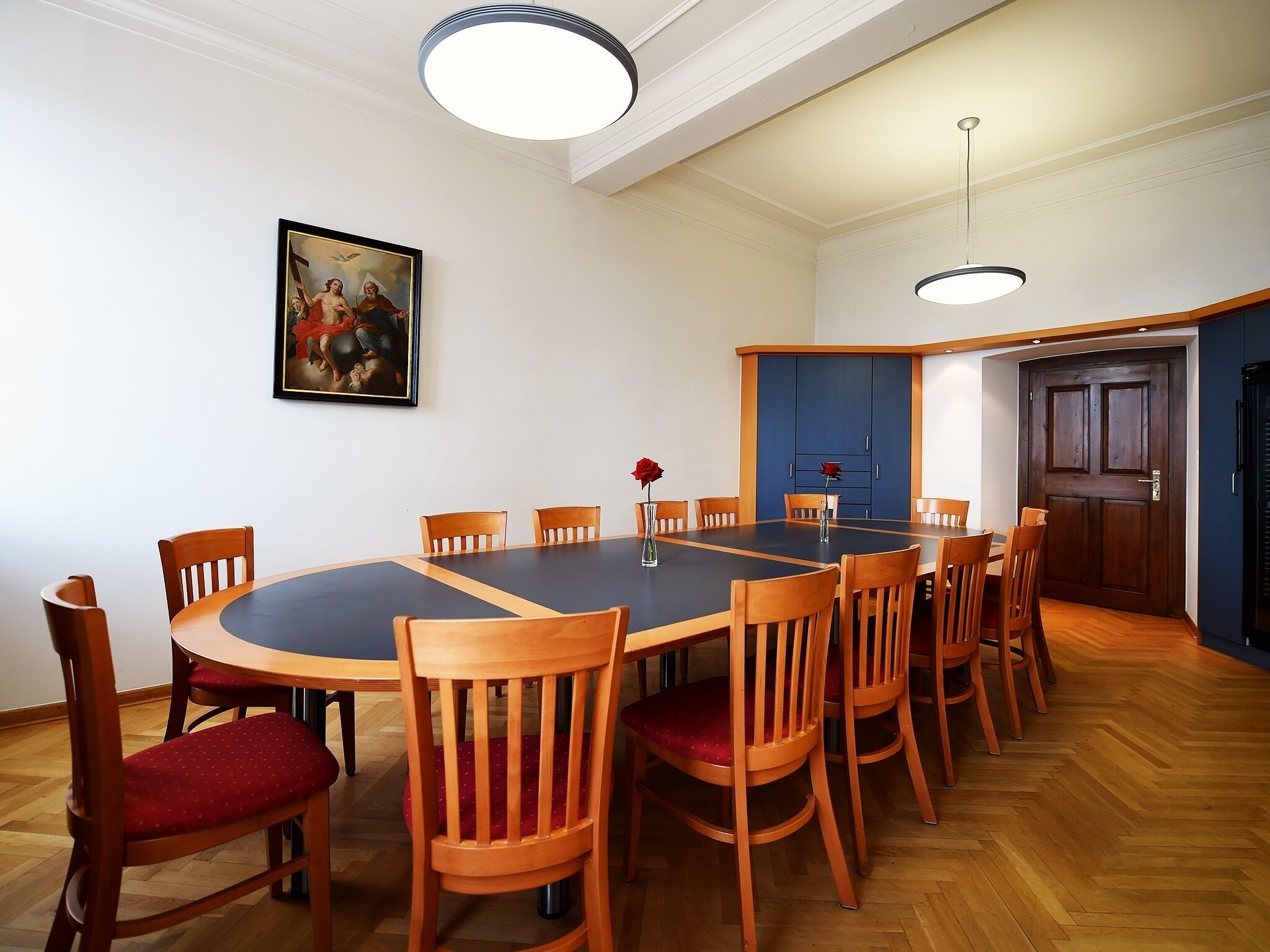 Un salón de conferencias con una mesa redonda, sillas de madera, un cuadro en la pared y una puerta que da a otra habitación.